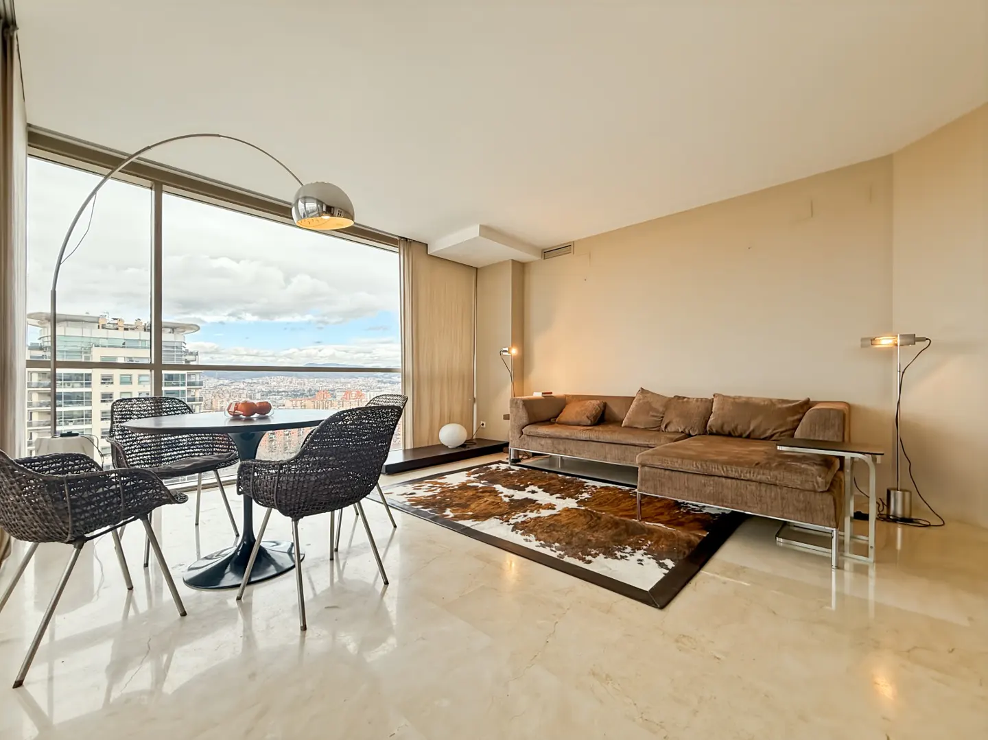 A modern living room with a brown sectional sofa, cowhide rug, and a round table with woven chairs. Large windows offer a city view.