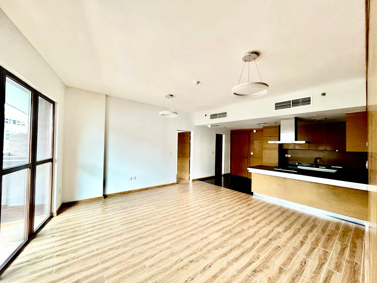 Bright, empty apartment with wood-look tile floors, white walls, and an open kitchen with dark countertops. Large windows let in natural light.