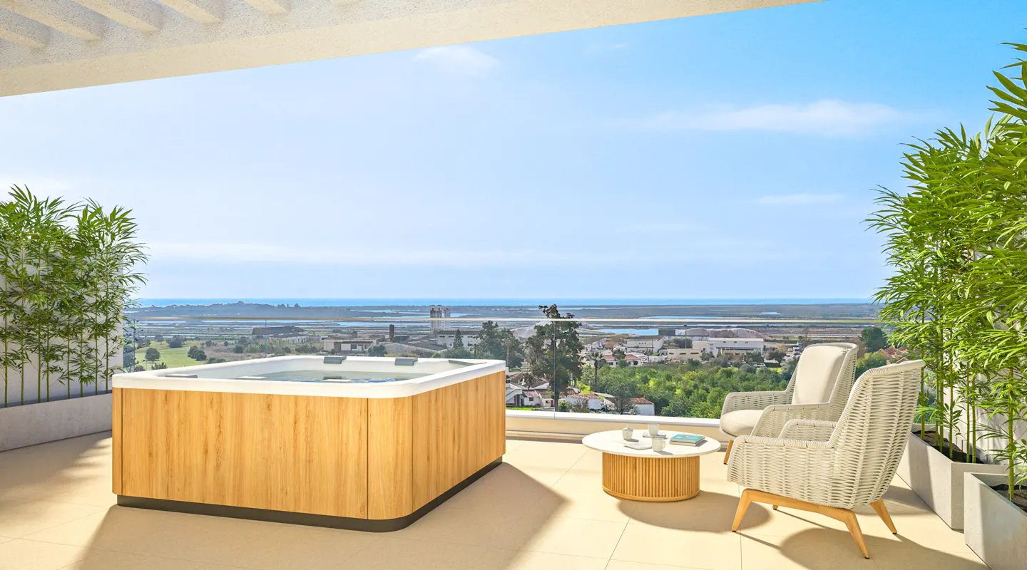 Balcony view with a wooden hot tub, two wicker chairs, and a small table overlooking a city and ocean under a blue sky.