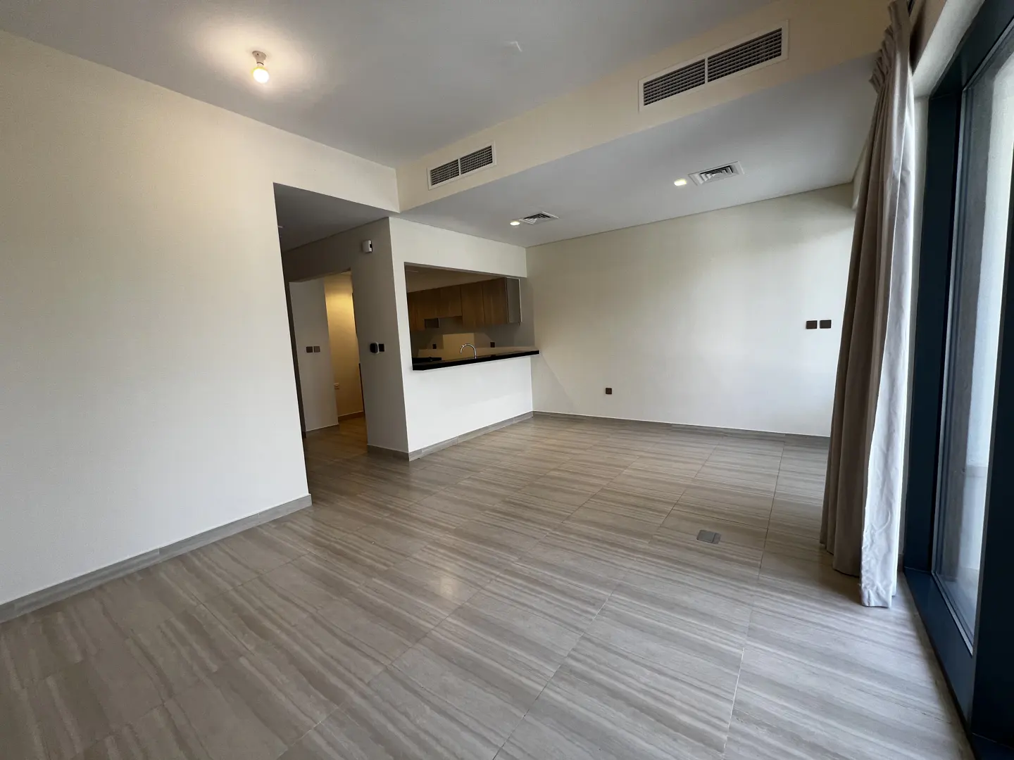 Unfurnished living room with gray tile floor, white walls, and open kitchen with wood cabinets. Large window with beige curtains.