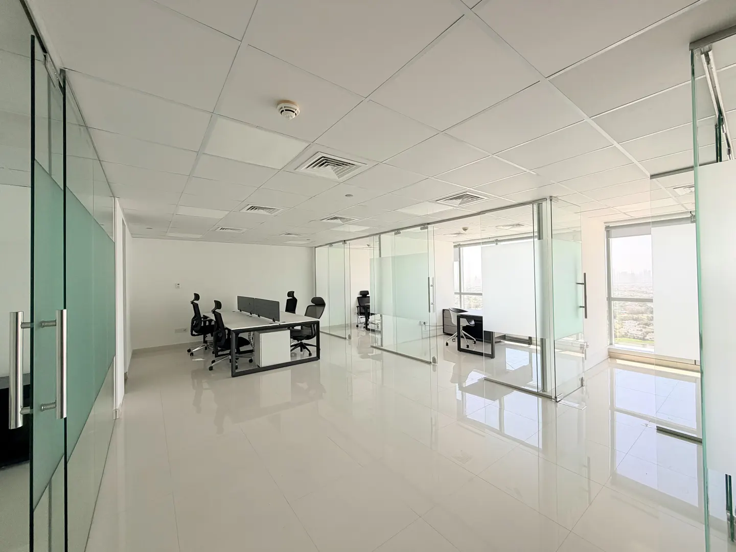 Bright, empty office space with glass-walled rooms, white tile floors, and desks with black chairs. Natural light streams through the windows.