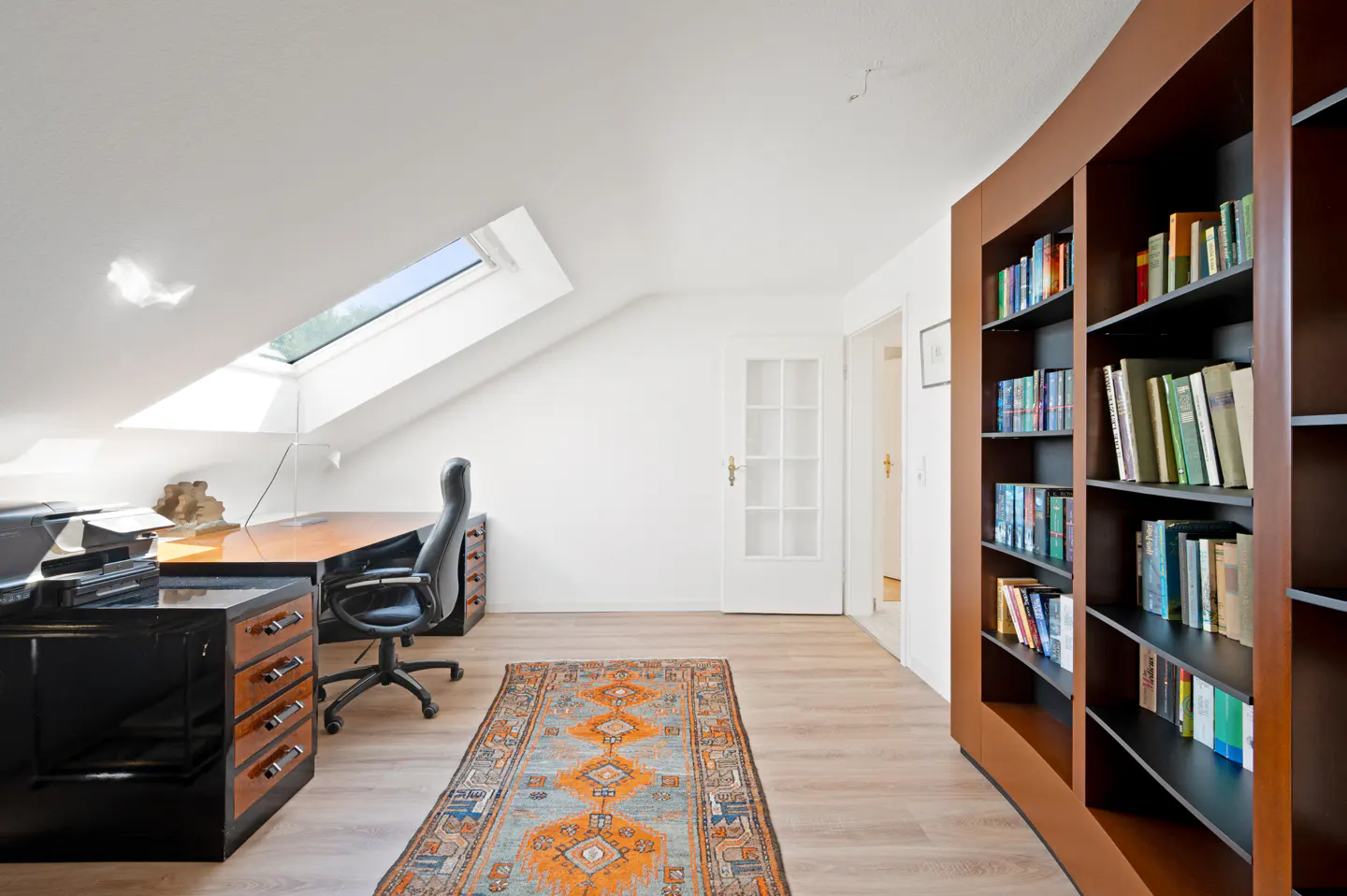 Attic office with a desk, black chair, and a brown bookcase filled with books. A patterned rug lies on the light wood floor.