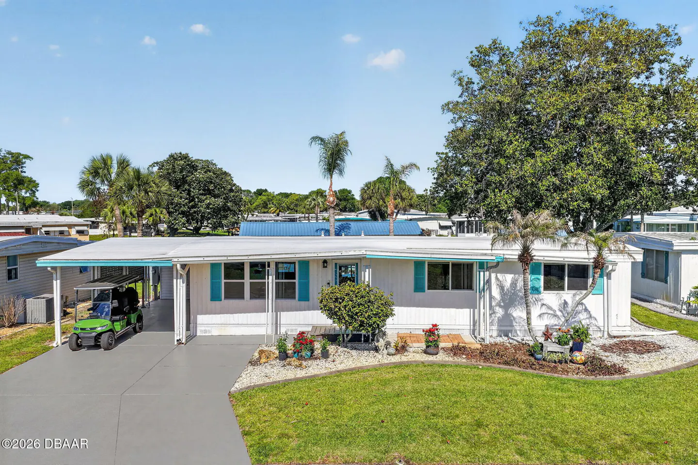 A white mobile home with turquoise shutters, a carport with a green golf cart, and a green lawn.