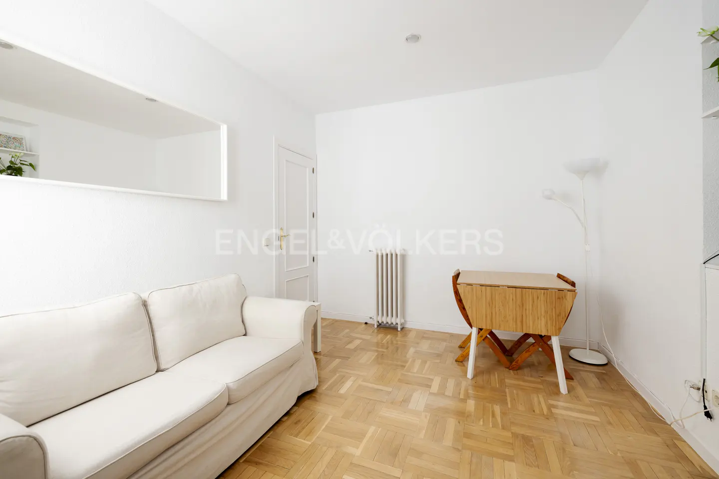 Bright living room with white walls, parquet floor, white sofa, wooden folding table, radiator, and a large mirror.