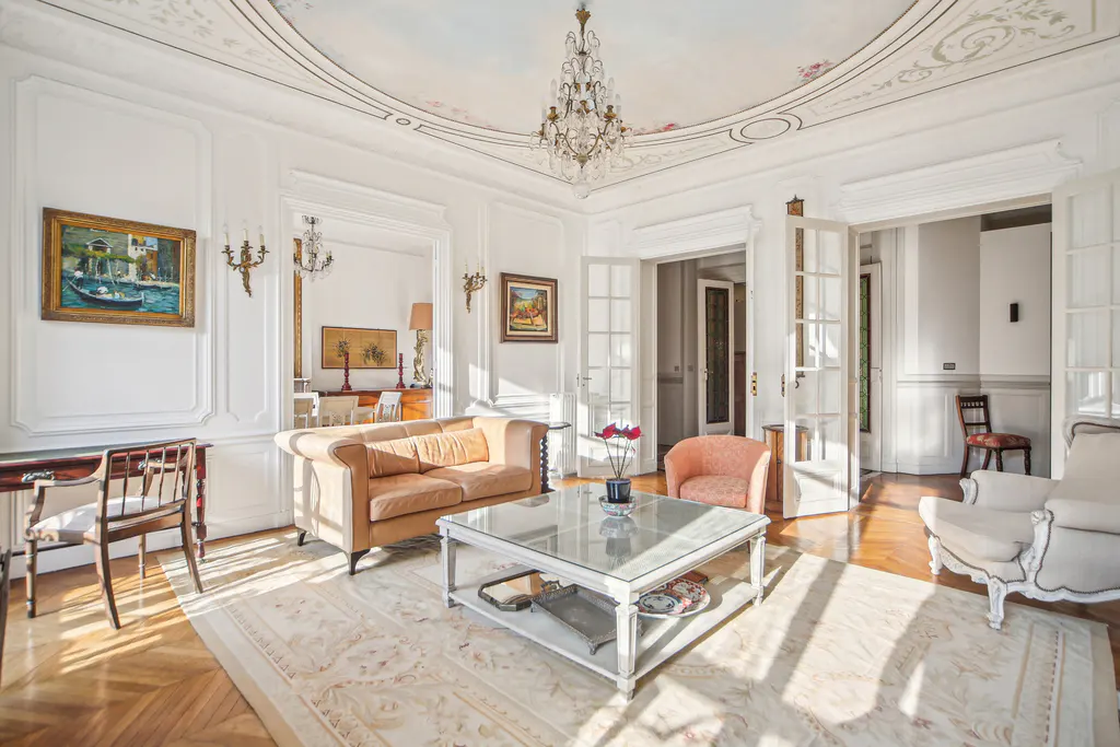 Elegant living room with a crystal chandelier, tan sofa, glass coffee table, and ornate white molding on the ceiling.
