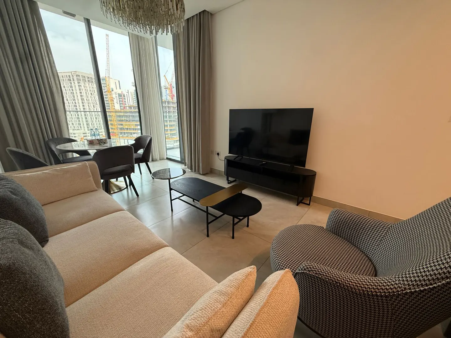 A modern living room with a beige sofa, houndstooth armchair, black TV stand, and a black coffee table. A round dining table sits by a large window with city views.