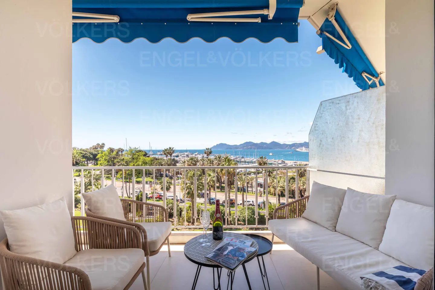 Balcony view with seating, table, and ocean vista. Blue awning above. Chairs, sofa, and table with wine and magazine. Palm trees and boats visible.