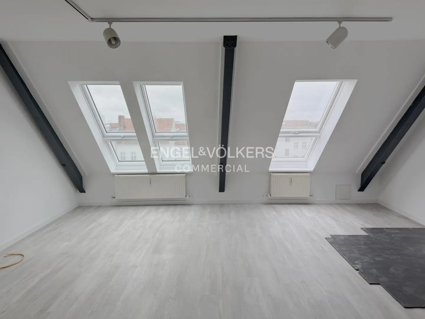 Bright attic room with white walls, gray wood-look floors, and two skylights. Black beams accent the ceiling. Radiators under the windows.