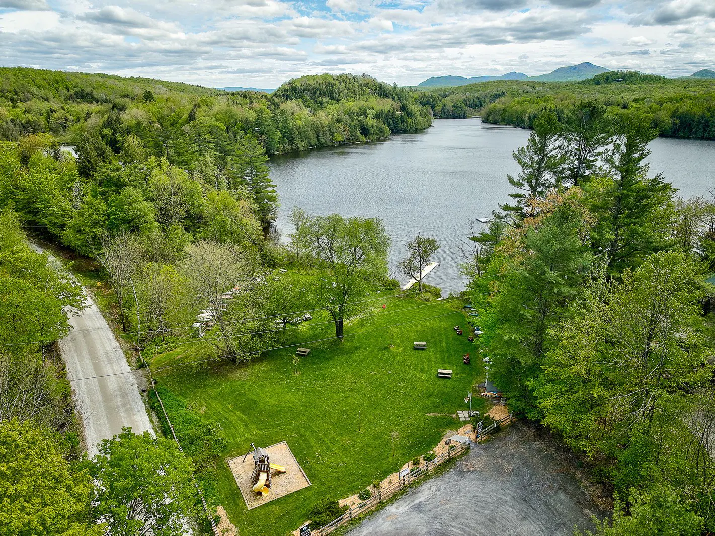 Aerial view of a grassy park with a playground and picnic tables, bordering a lake surrounded by lush green forests.