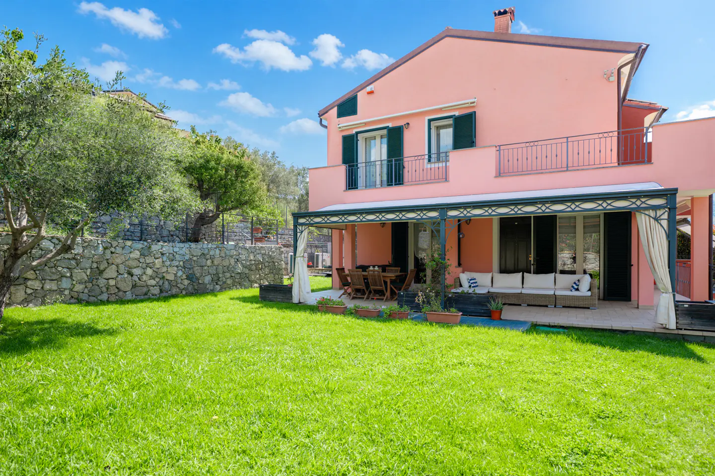 Two-story pink house with green shutters, a balcony, and a covered patio with furniture, surrounded by a green lawn and stone wall.