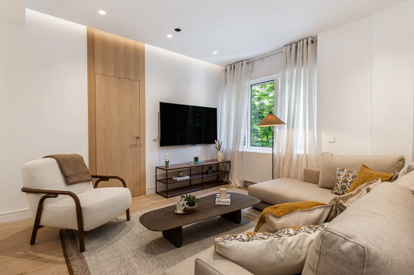 Living room with a beige sectional sofa, a white armchair, a dark wood coffee table, and a TV on the wall.