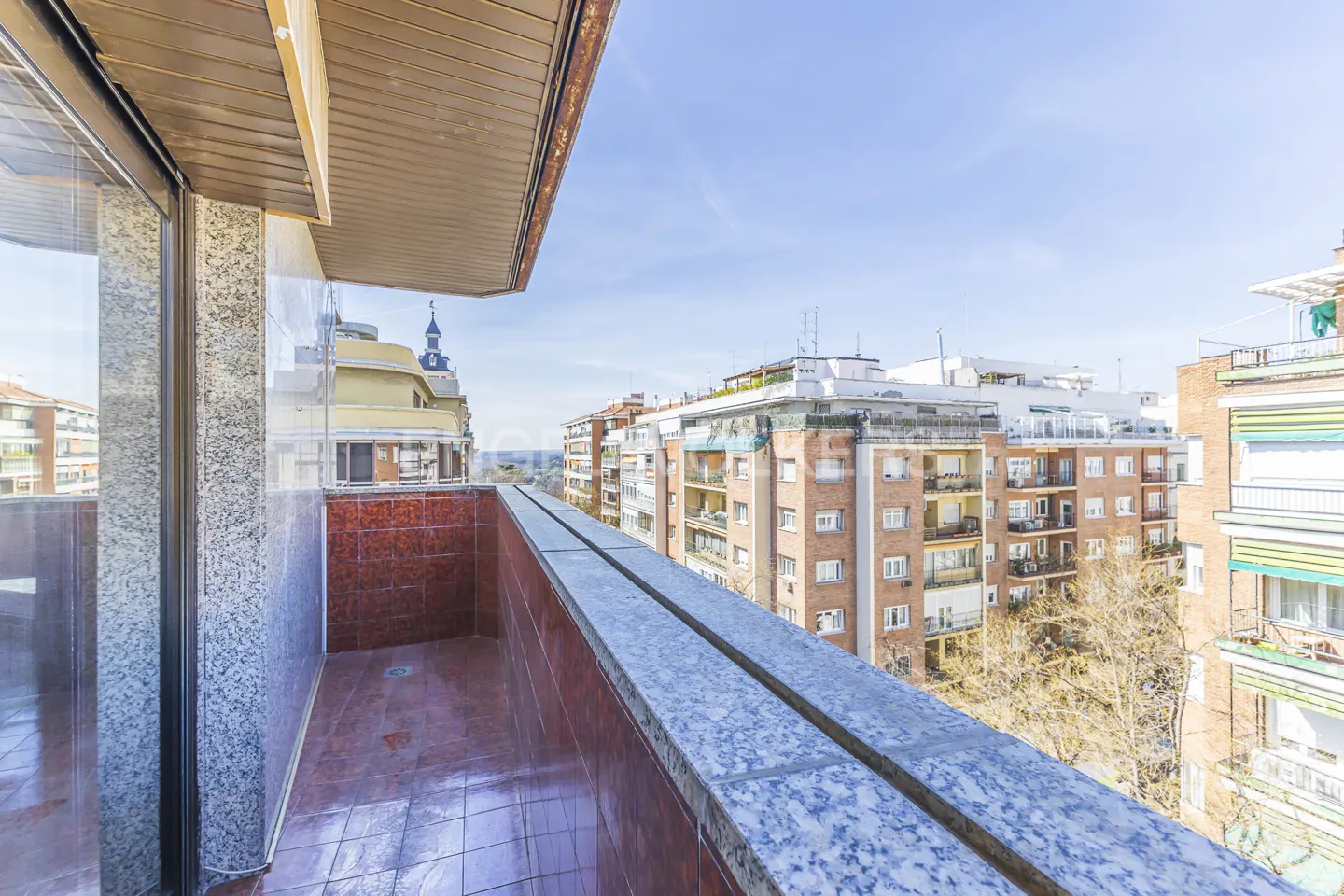View from a balcony with red tile floor and marble railing, overlooking city buildings under a blue sky.