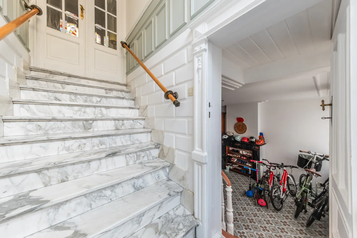 Interior view of a home's entrance with marble stairs, a wooden handrail, and a doorway leading to a tiled area with bikes and storage.
