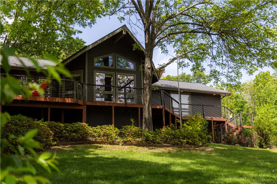 A two-story gray house with a deck and large windows is surrounded by green trees and grass.