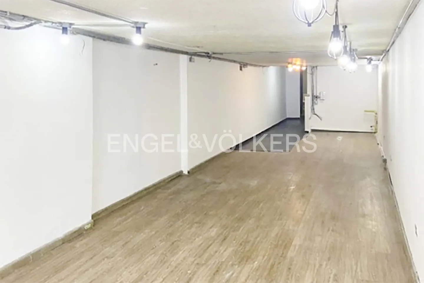Empty commercial space with white walls, wood floors, and exposed lighting. Engel & Völkers logo on the wall.