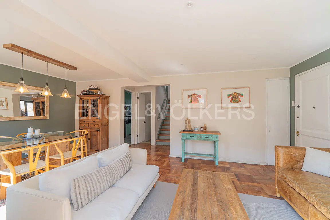 Living room with a white sofa, a wooden table, and a glass-top dining table with yellow chairs. There is a staircase in the background.