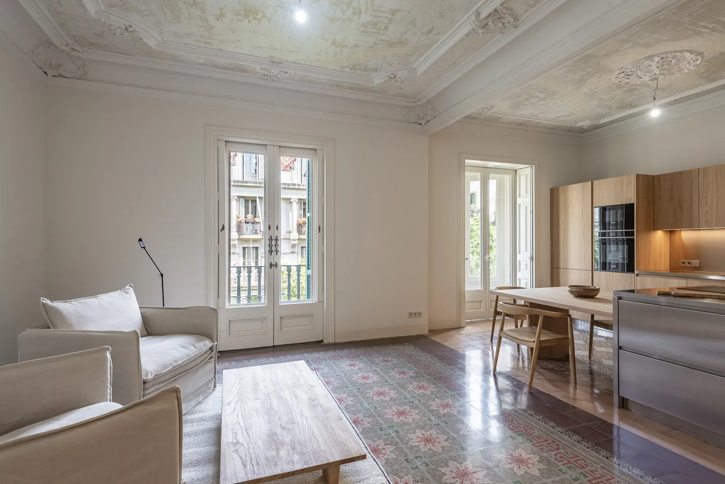 Bright, open-concept apartment with white walls, ornate ceiling, and patterned tile floor. Cream armchairs face a wooden coffee table. Kitchen with wood cabinets visible.