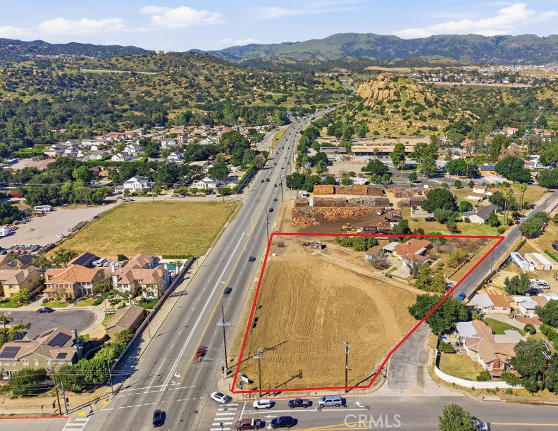 Aerial view of a vacant lot outlined in red, adjacent to a busy road with cars, houses, and green hills in the background.