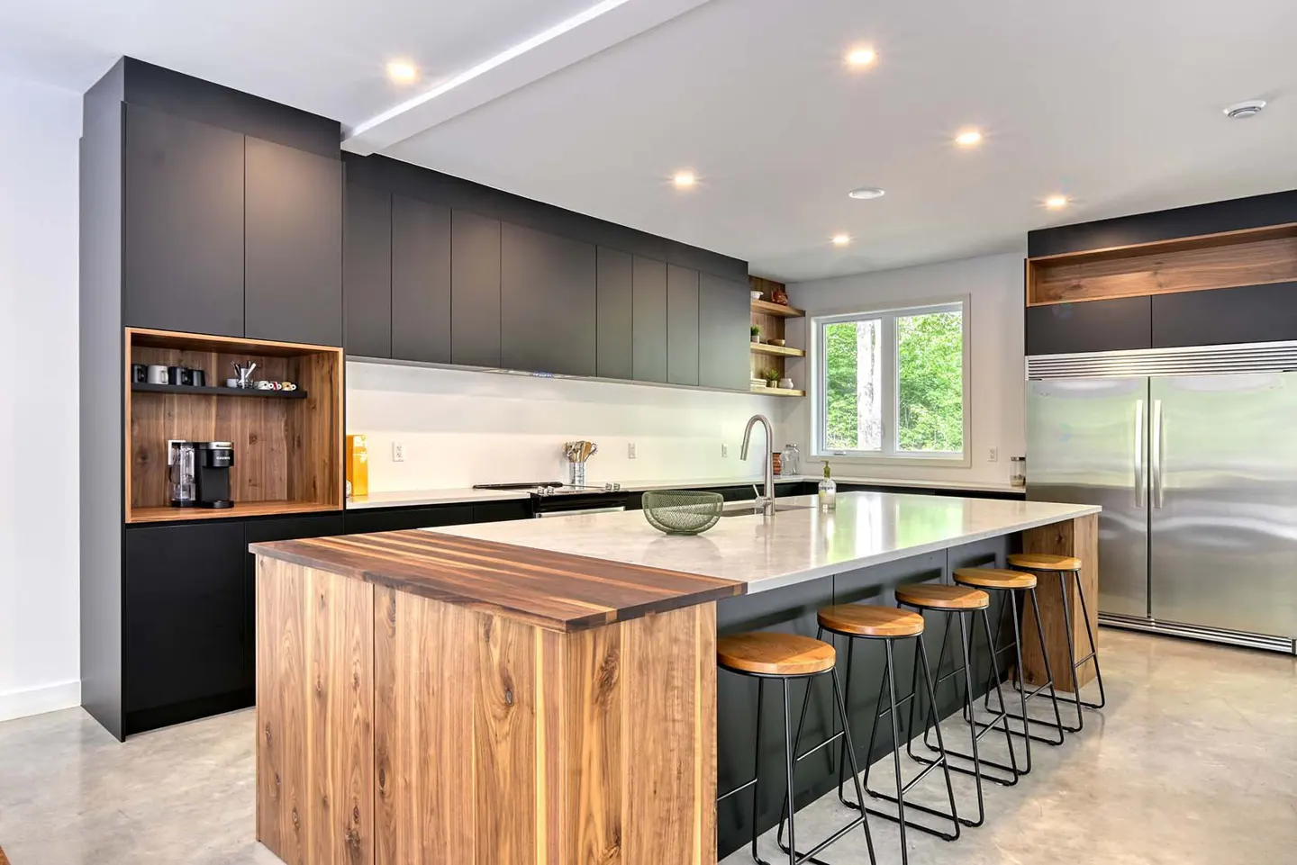A modern kitchen with black cabinets, a wood-paneled island, and white countertops. Stools line the island, and a stainless steel refrigerator is visible.
