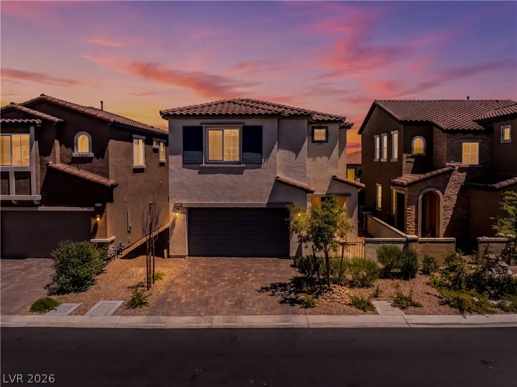 Two-story stucco house with a dark garage door and brick driveway at dusk. The sky is pink and purple.