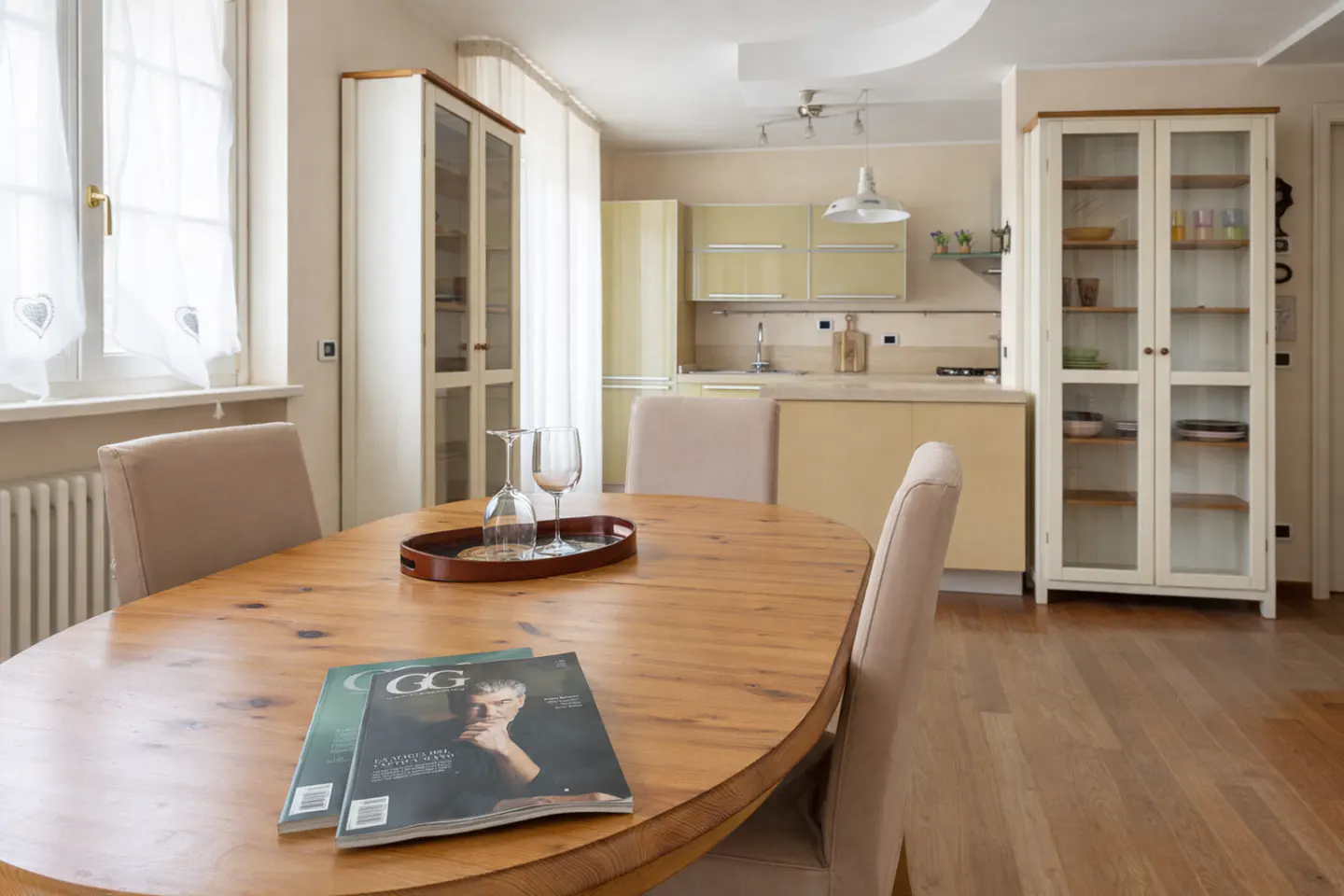 Interior view of a dining area with a wooden table, chairs, magazines, and wine glasses. A kitchen and cabinets are in the background.