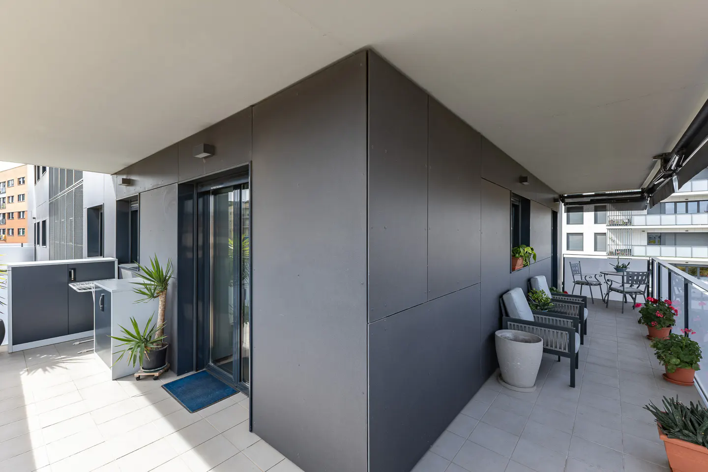Modern apartment balcony with dark grey walls, tiled floor, outdoor seating, and potted plants under a covered ceiling.