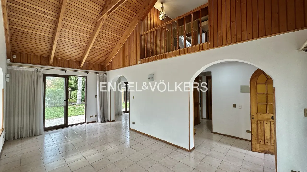Interior view of a home with a wood ceiling, white walls, tile floors, and a balcony overlooking the living space. Sliding glass doors lead to a green yard.