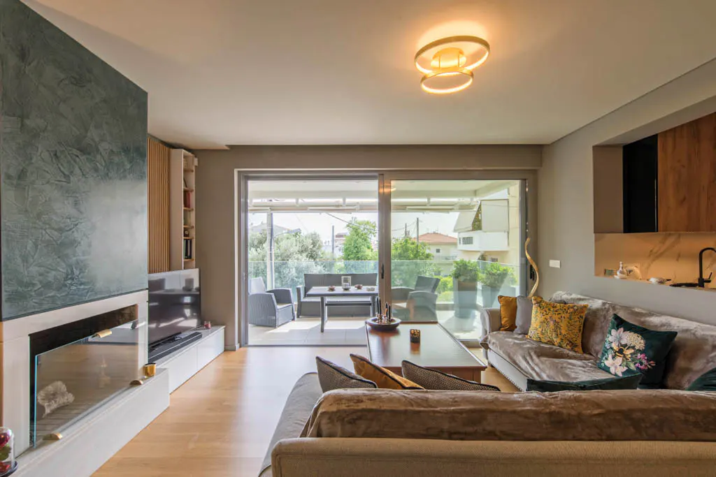 Living room with gray sofas, decorative pillows, and a coffee table. A fireplace and TV are on the left, and a balcony with outdoor furniture is visible through sliding glass doors.