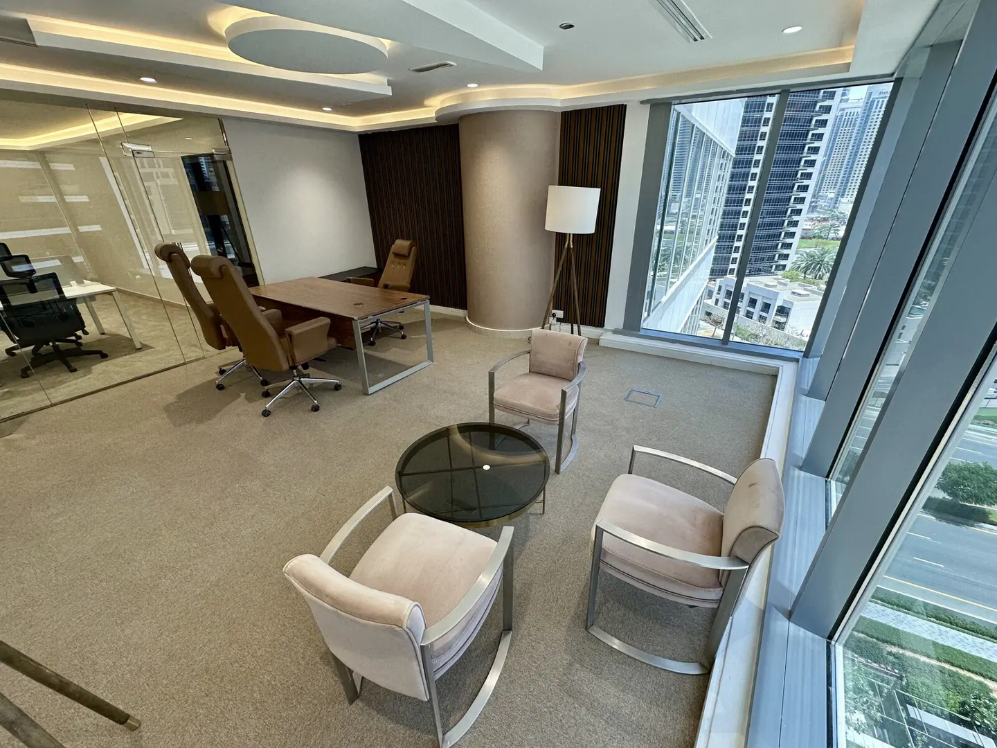 Office space with a brown desk, tan chairs, and a black coffee table. Large windows offer a city view.