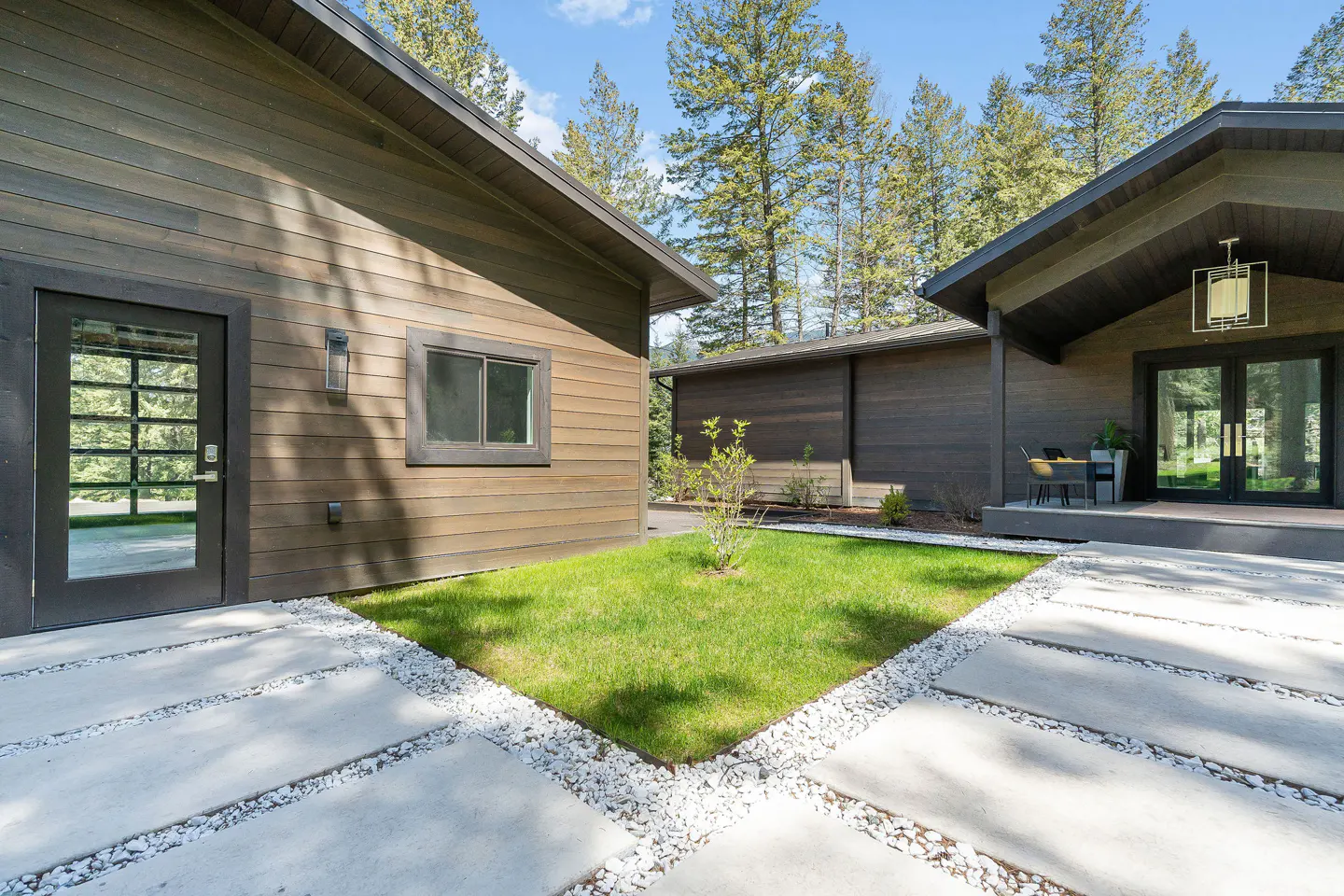 Exterior view of a modern brown house with a concrete patio and green lawn. Trees are visible in the background.