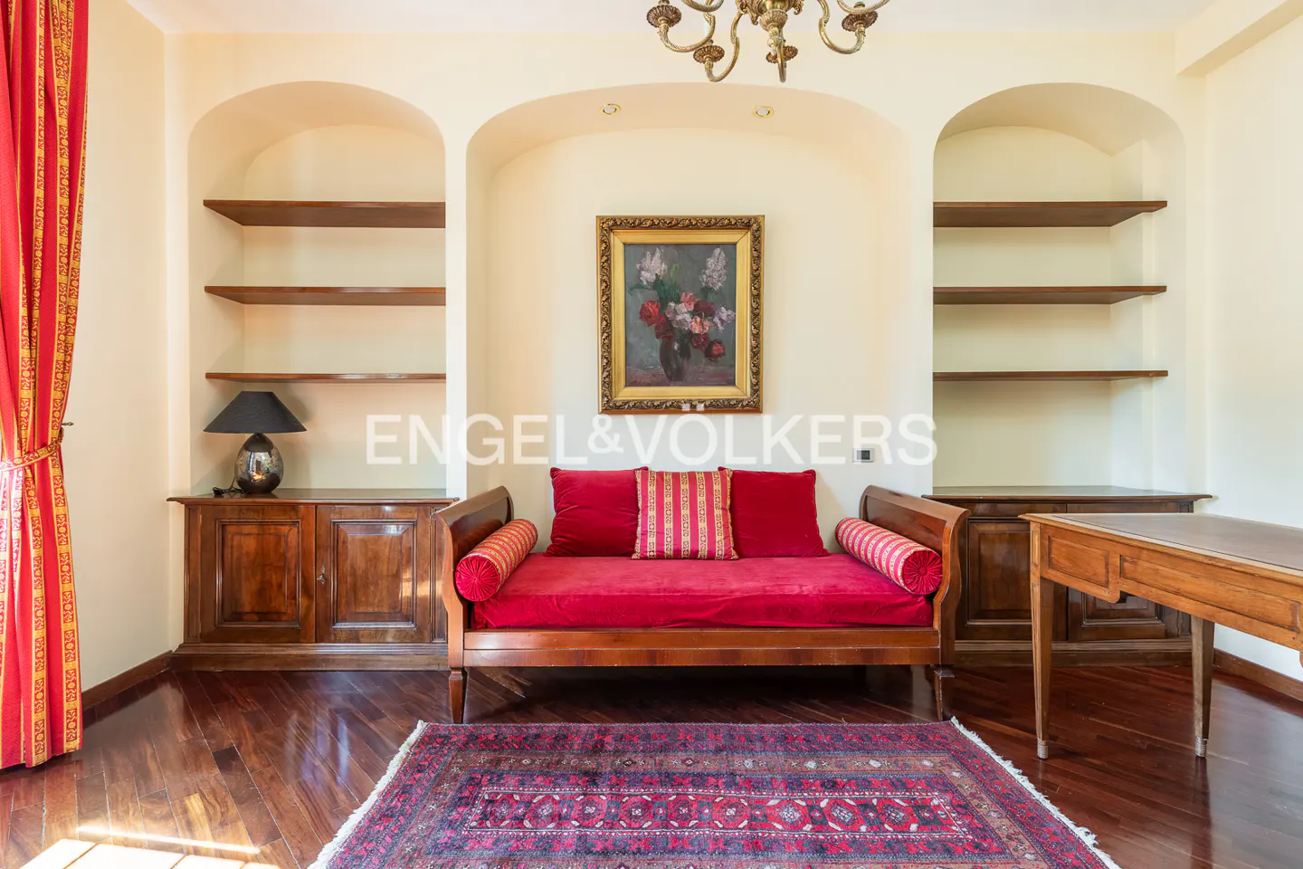 A living room with a red daybed, wooden floors, and built-in shelves. A painting hangs above the daybed.