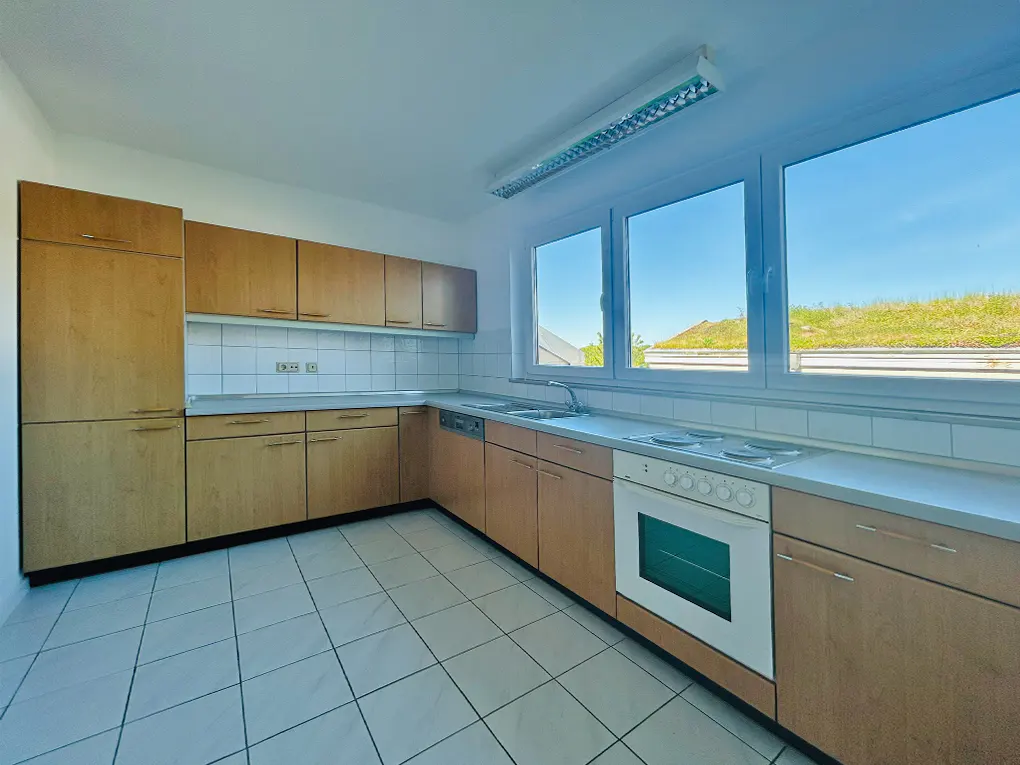 Bright kitchen with light wood cabinets, white tile floor and backsplash, and a white oven. A window shows a green roof outside.