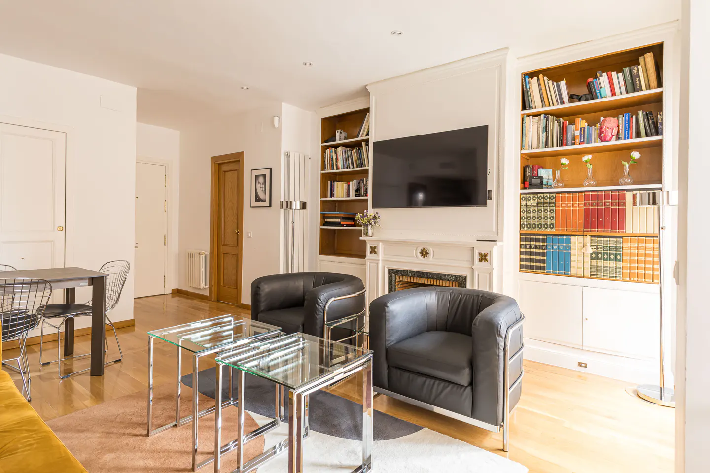 Living room with two black leather chairs, glass tables, a TV over a fireplace, and built-in bookshelves filled with books.