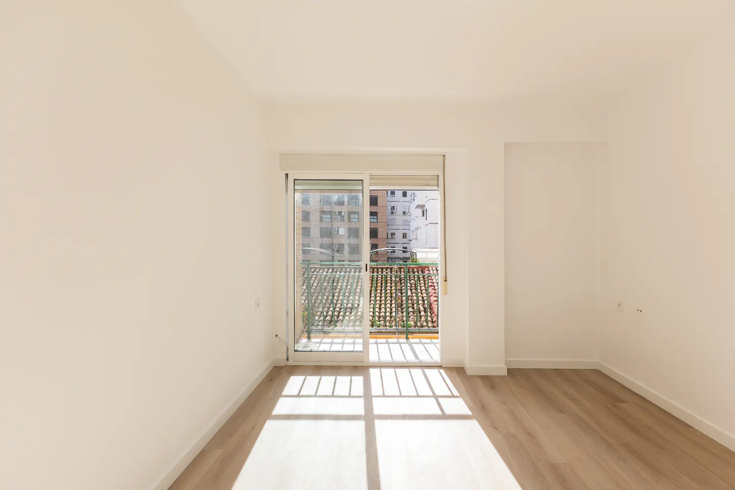 Bright, empty room with white walls and light wood floors. Sliding glass doors open to a balcony with a view of buildings. Sunlight streams in.