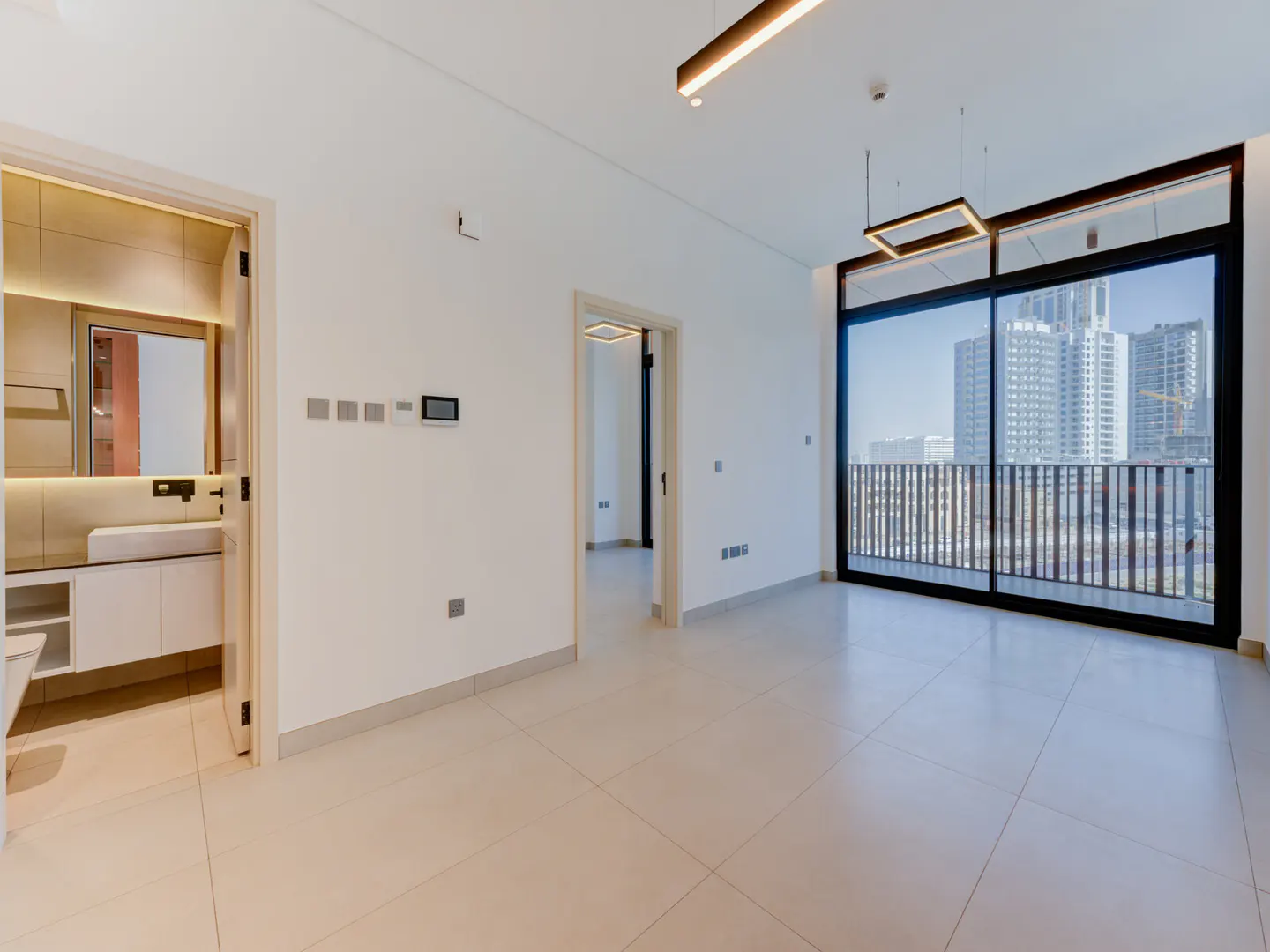 Bright, empty room with beige tile floors, white walls, and a large window with a city view. A bathroom is visible through an open door.