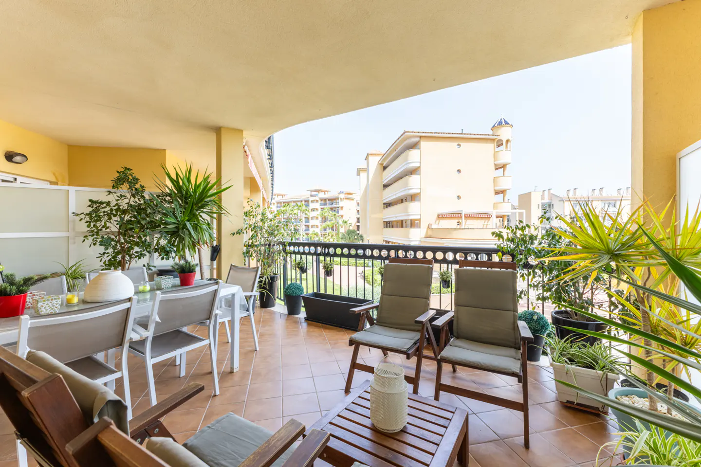 A sunny balcony with outdoor furniture, plants, and a view of nearby buildings.