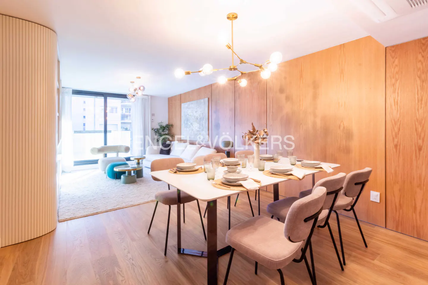 A bright, modern dining area with a set table, chairs, and a gold chandelier. A living room with a white sofa is visible in the background.