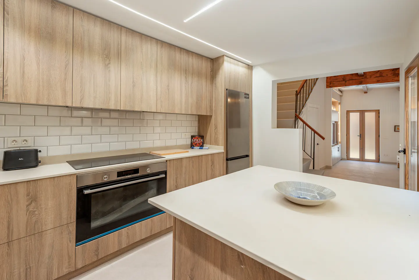 Modern kitchen with light wood cabinets, white countertops, stainless steel appliances, and a white island with a decorative bowl. Staircase in background.