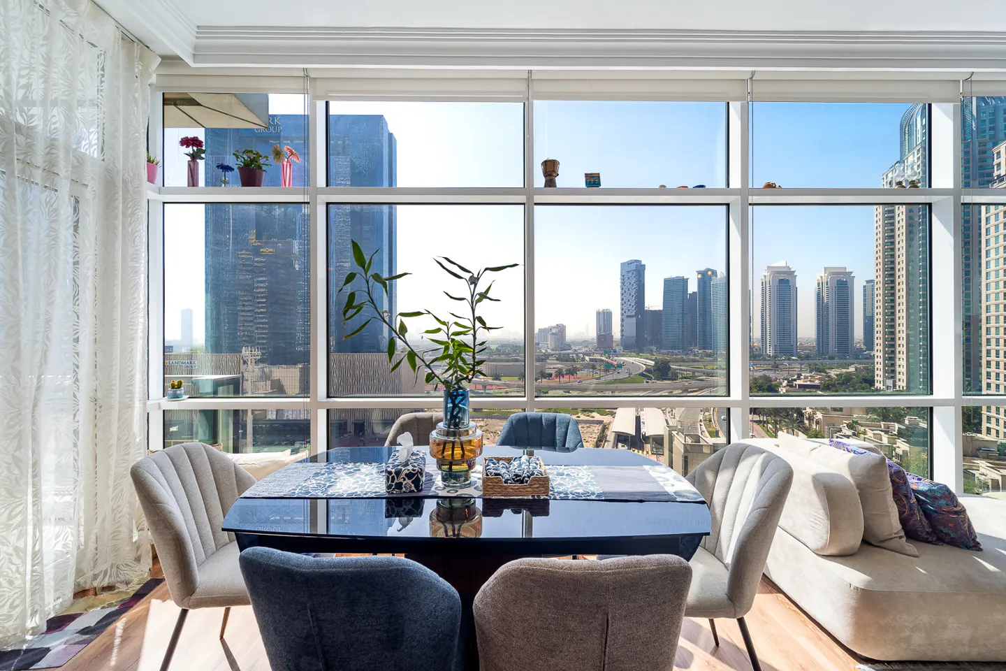 Dining area with a black table, chairs, and a large window overlooking a city skyline. A beige chaise lounge is on the right.