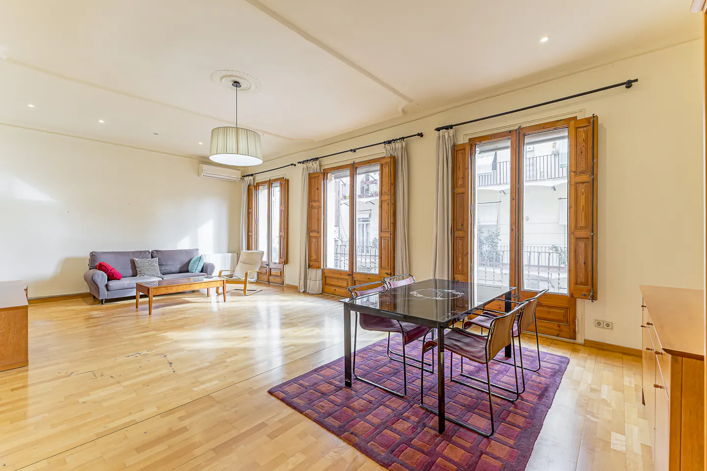 Bright living room with wood floors, a gray sofa, and a dining table with four chairs on a purple rug. Large windows with wood shutters.