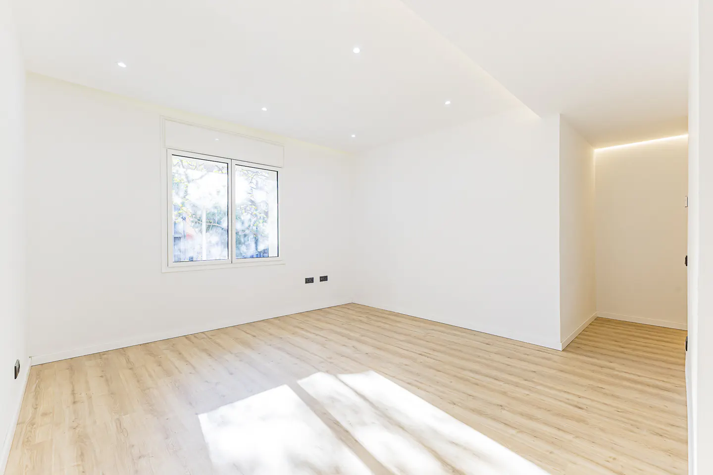 Bright, empty room with light wood floors, white walls, and a window. Recessed lighting in the ceiling. Hallway visible on the right.