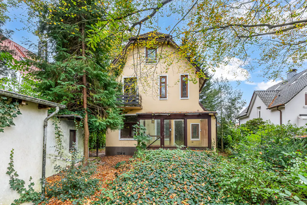 Back view of a two-story beige house with a glass-enclosed porch and a small balcony, surrounded by lush greenery.