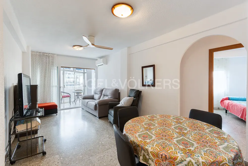 Bright living room with a dining table, grey sofa, armchair, TV, and a balcony with outdoor furniture.