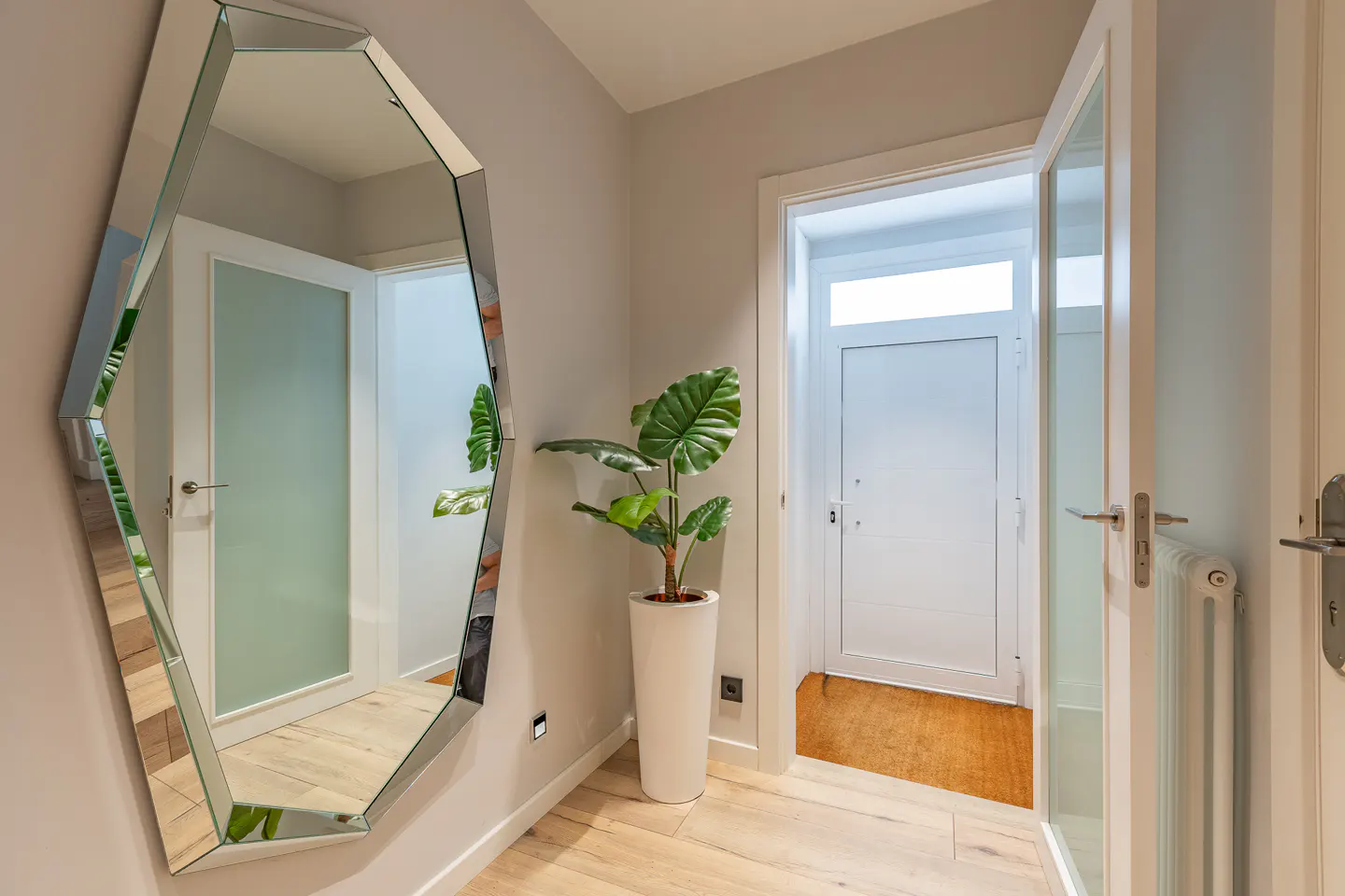 Hallway with a large mirror, potted plant, and open white door. The floor is light wood.