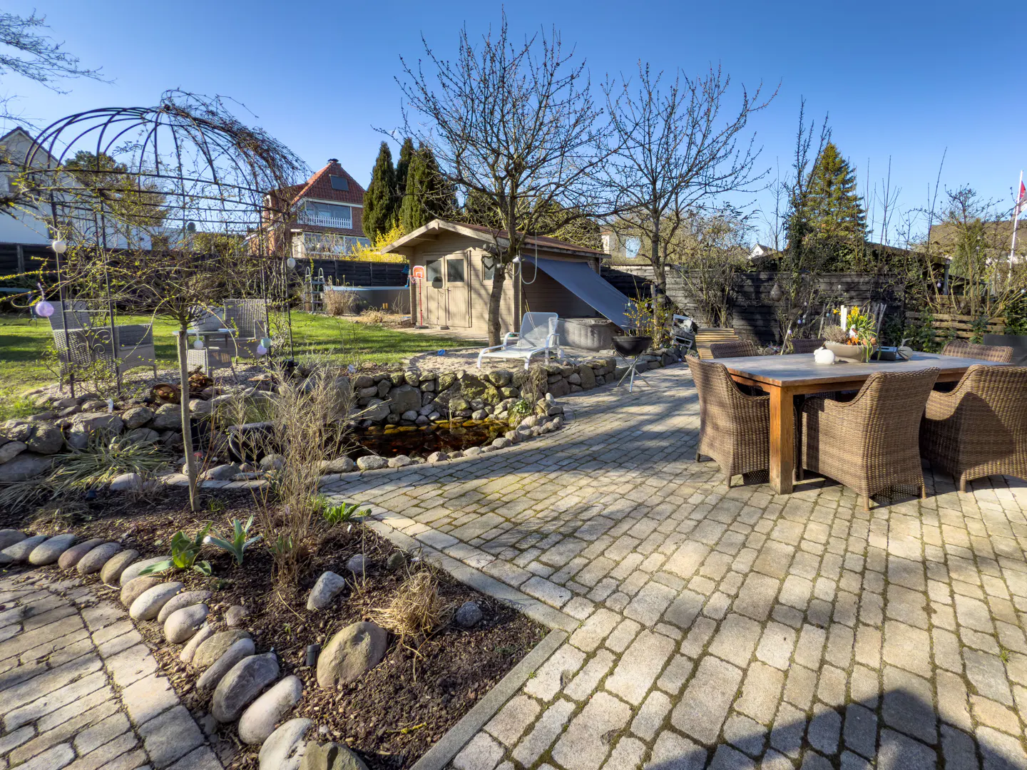 Outdoor patio with stone pavers, a dining table with wicker chairs, a pond, and a small wooden shed in the background.