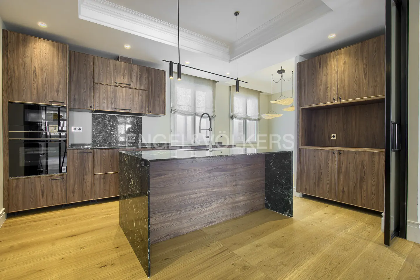A modern kitchen with wood cabinets, marble countertops, and a large island with a sink. Light wood floors and natural light fill the space.