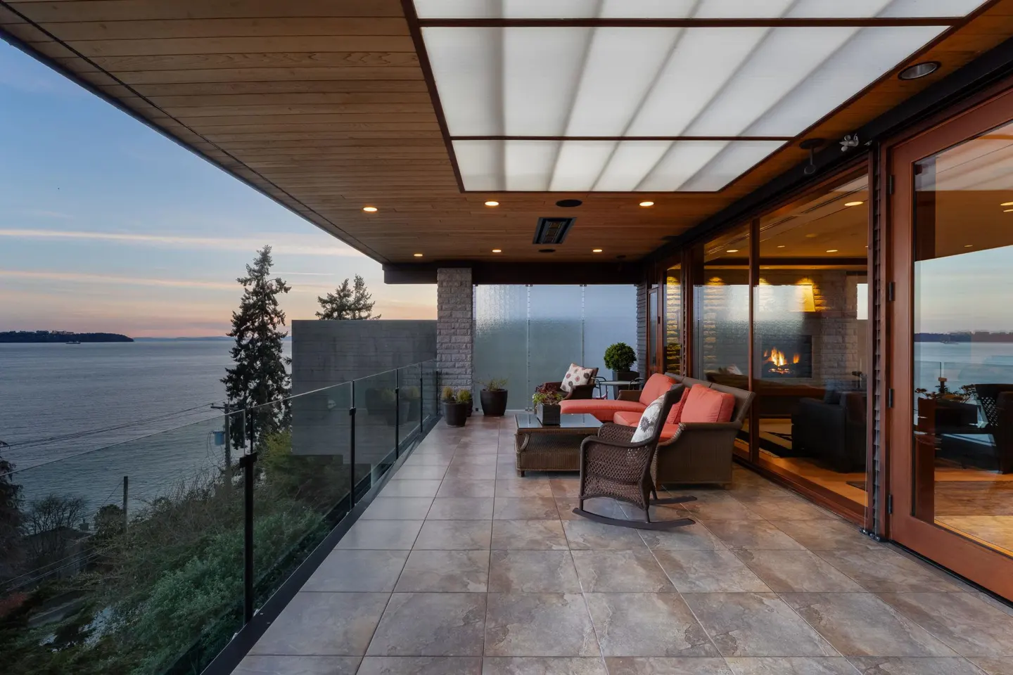 Covered patio with orange sofa, wicker rocking chair, and glass railing overlooking the ocean at dusk.