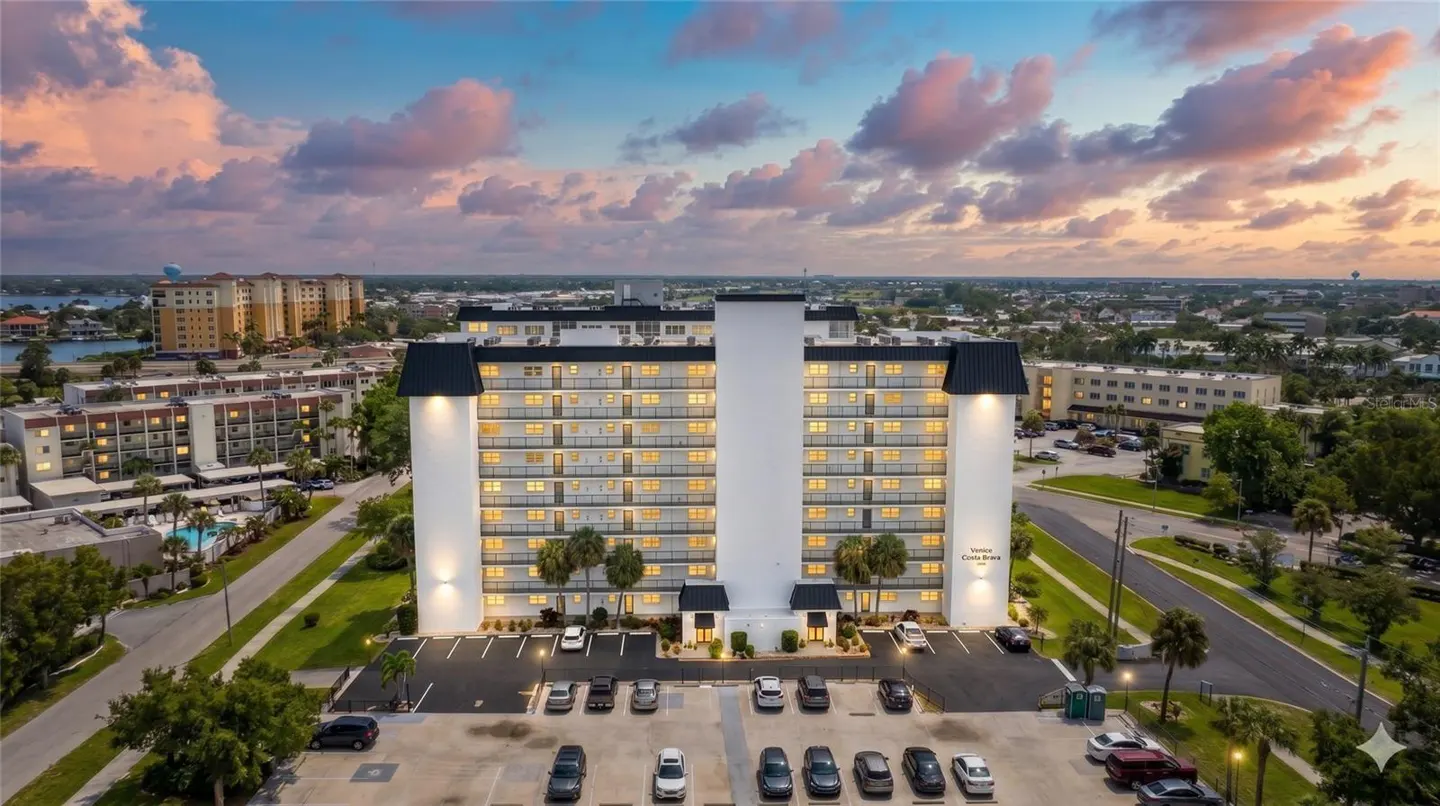Aerial view of the white Harbor Crosse condo building with black roofs, a parking lot, and a sunset sky.