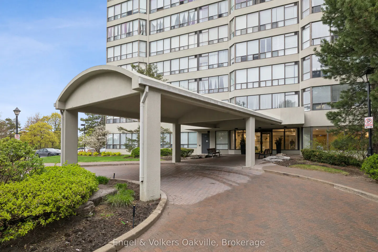 Exterior view of a high-rise apartment building with a covered entrance. The entrance has a brick driveway and manicured landscaping.