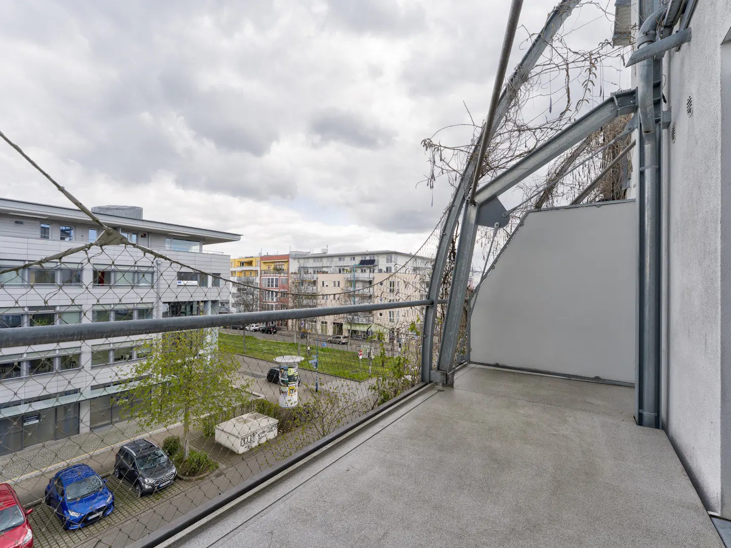 View from a balcony with metal railings overlooking a street with parked cars and buildings under a cloudy sky.
